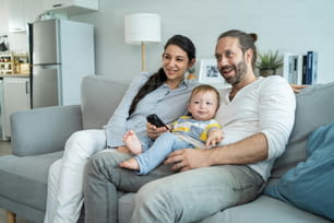 自宅のリビングルームで赤ん坊の子供と映画を見ている白人のカップル。ソファに座ってリモコンのテレビ番組を使用し、若い息子を抱きながらテレビを見て笑うのを楽しんでいる美しい幸せな家族