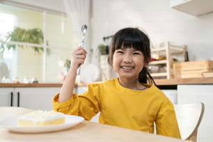 Porträt eines asiatischen glücklichen jungen Kindes, das zu Hause in der Küche Kuchen isst. Entzückendes kleines Kind sitzt auf dem Tisch und fühlt sich glücklich und genießt süßes Essen nach dem Kochen von Speisen oder dem Backen von Hefeteig im Haus.