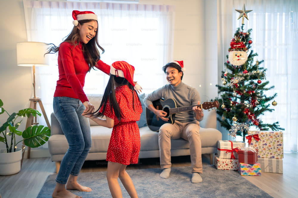 Un encantador miembro de la familia asiática disfruta cantando canciones navideñas juntos en casa. La pequeña hija se siente feliz y emocionada de celebrar la fiesta de Acción de Gracias navideña junto con los padres en casa.