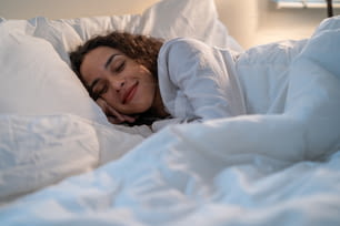 暗い夜の部屋の寝室のベッドに横たわっているラテン系の美しい女性。パジャマを着た魅力的な若い女性が、自宅での健康管理のための静かな部屋で快適な枕と居心地の良い毛布で一人で寝ています。