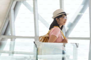 Mulher asiática na camisa rosa usar óculos, chapéu com mochila amarela está esperando o voo no hall do aeroporto.