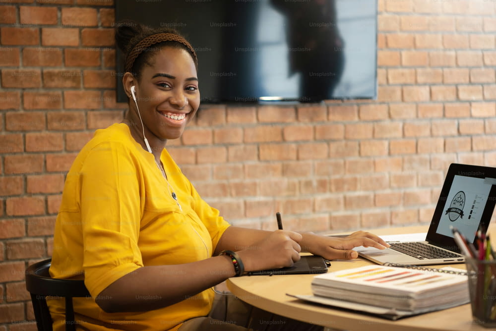 Porträt von African FeMale Creative Designer in gelbem Hemd mit Kopfhörern sitzend während der Verwendung von Grafik-Tablet-Arbeit auf Laptop-Computer.