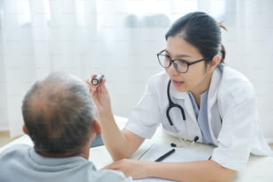 Junge asiatische Ärztinnen tragen eine Brille, die die Augen älterer Patienten mit Taschenlampe in der Arztpraxis überprüft.