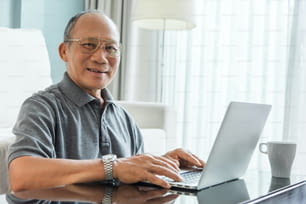 Relaxed Asian Senior man using laptop at his house. Happy Elderly Male laughing. Looking camera. Retirement