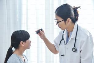 젊은 아시아 여성 의사는 의료 사무실에서 손전등으로 어린 소녀 환자의 눈을 확인하는 안경을 착용합니다.