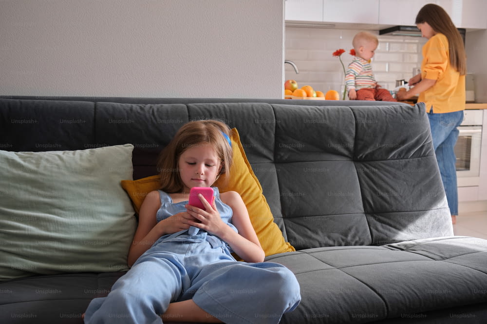 Una bambina che si siede su un divano che guarda un telefono cellulare