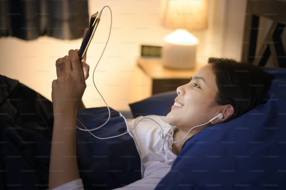 Una mujer joven está usando una tableta, viendo películas o videollamadas a sus amigos o familiares en su dormitorio, luz nocturna