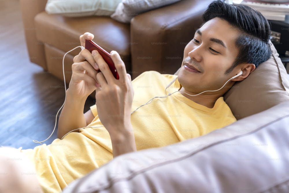 jeune main masculine asiatique jouant à des jeux sur smartphone en ligne occasionnel et de loisirs sortant et jpyful allongé sur le canapé le week-end à la maison concept d’idées d’activités de quarantaine