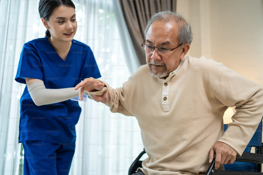 Asian Caregiver girl doctor support older disabled man walk in house. Attractive nurse taking care of mature senior elderly male patient sitting on wheelchair at home nursing care and doing therapy.
