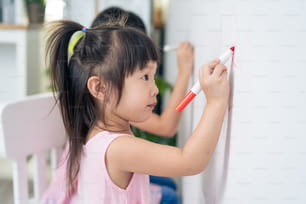 Asiatische junge Geschwister Kind Mädchen genießen Farbe auf weiße Wand im Wohnzimmer. Kleine entzückende Kinder, die Spaß beim Zeichnen und Ausmalen von Kunstbildern mit Hapiness haben, genießen kreative Aktivitäten im Urlaub zu Hause.