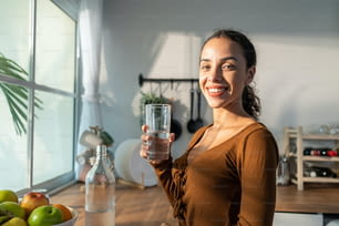 Giovane bella donna latina che tiene l'acqua pulita in vetro in cucina. Attraente ragazza assetata attiva bere o prendere un sorso di minerale naturale in tazza per la cura della salute e il benessere in cucina in casa.