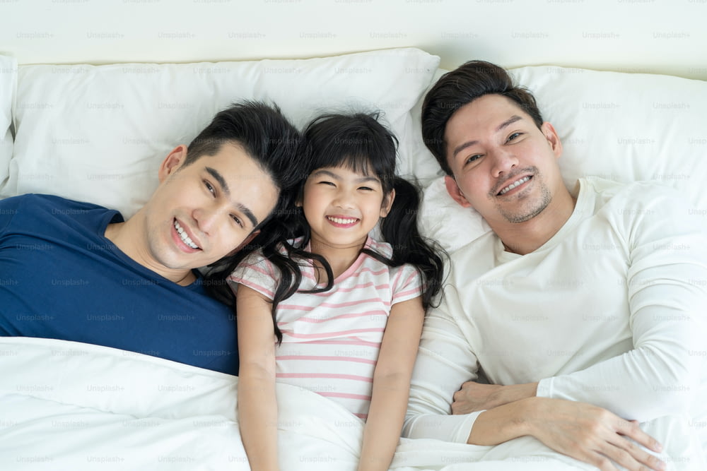 Retrato de una familia gay de hombre guapo con una hija pequeña en el dormitorio. Atractiva pareja romántica lgbt masculina acostada en la cama con una niña adorable por la mañana y mirando a la cámara en la casa