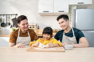 Une famille gay LGBTQ attrayante asiatique enseigne à une fille à faire de la pâte à levure. Beau couple d’hommes prendre soin et passer du temps avec un petit enfant adorable cuire la boulangerie dans la cuisine, profiter de l’activité parentale à la maison.