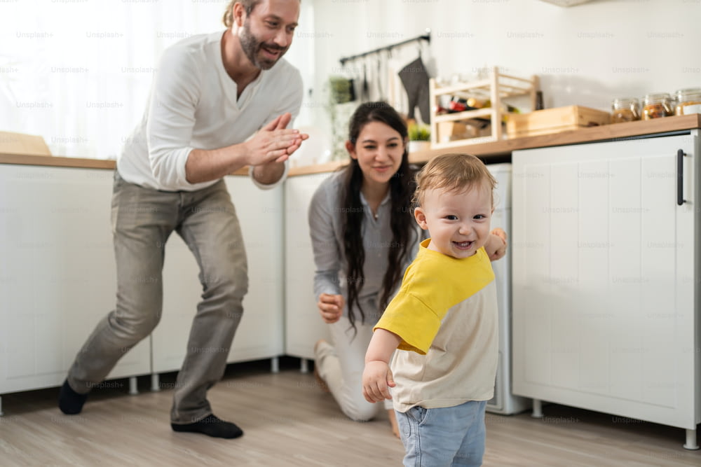 白人の男の子の赤ちゃんは、家で両親のサポートを受けて歩くことを学びます。幸せな家族、母親、父親は、幼い息子が自宅のリビングルームでスキルを伸ばすために床を歩くのを手伝います。