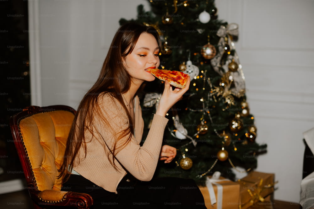 Una mujer comiendo una rebanada de pizza frente a un árbol de Navidad