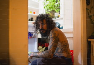 Un uomo con capelli lunghi e tatuaggi seduto davanti a un frigorifero