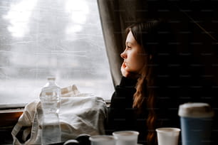 Una mujer mirando por una ventana con tazas sobre una mesa
