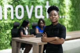 Jovem empresária africana confiante de pé com os braços cruzados em uma sala de reuniões de escritório com colegas trabalhando ao fundo