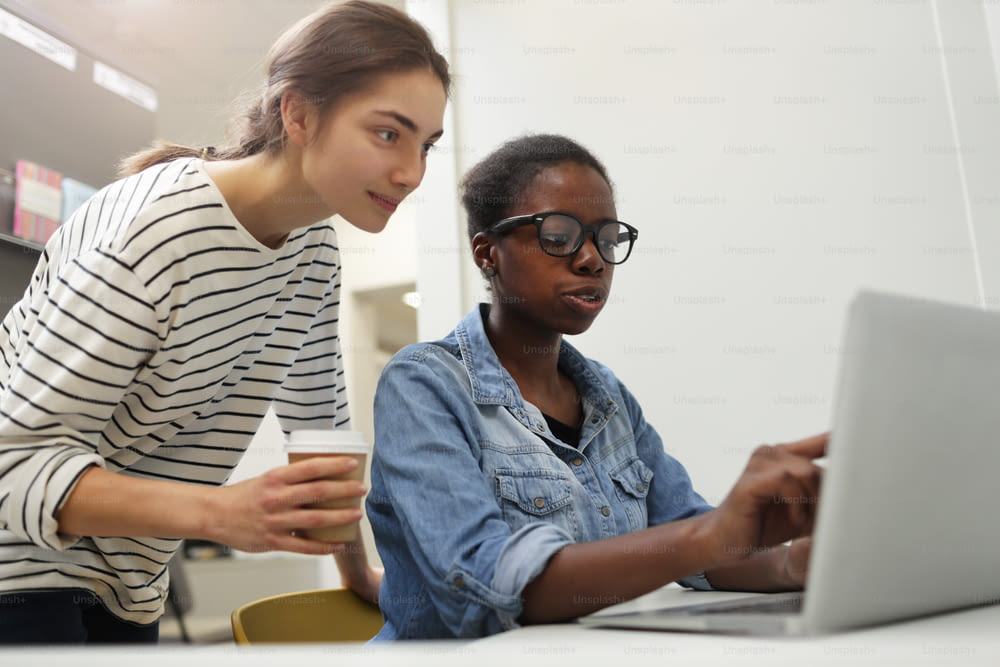 African young woman in eyeglasses sitting at the table and pointing at laptop she discussing online work with her colleague