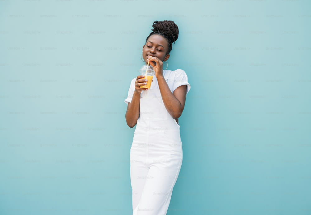 Young woman in white clothes drinking orange juice while standing at blue wall