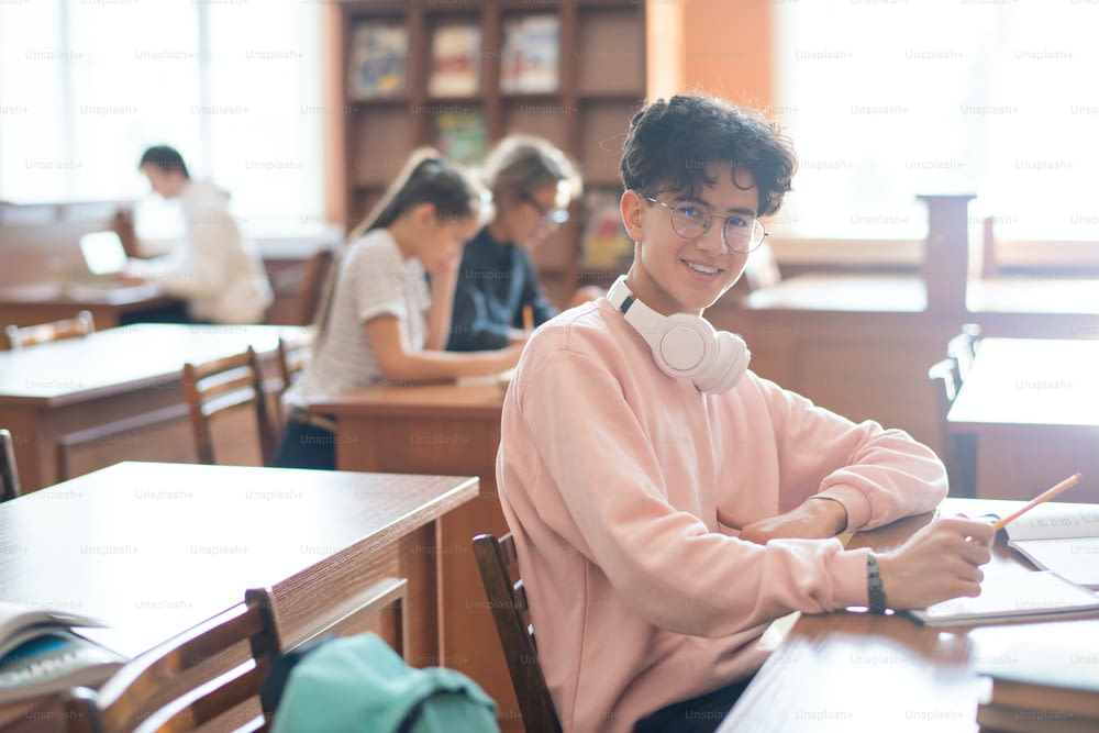 Smiling teenager in eyeglasses and casualwear looking at you while sitting by desk in college library and studying