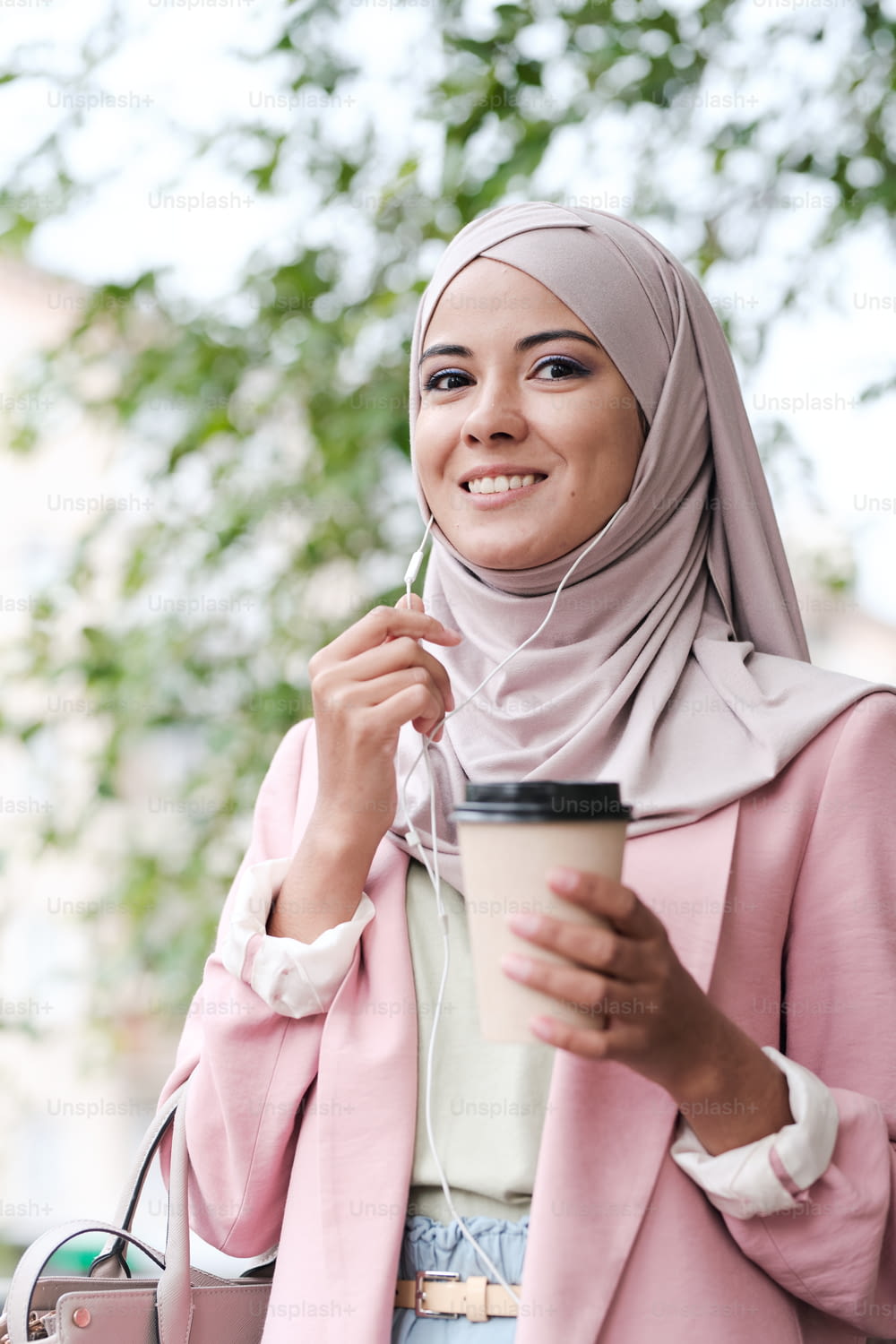 Young gorgeous smiling Muslim woman with glass of coffee talking to someone on the phone while standing in urban environment