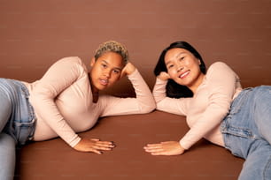 Zwei junge zeitgenössische Frauen verschiedener Ethnien in Blue Jeans und weißen Pullovern, die Sie mit einem zahnigen Lächeln ansehen, während Sie auf dem Boden liegen