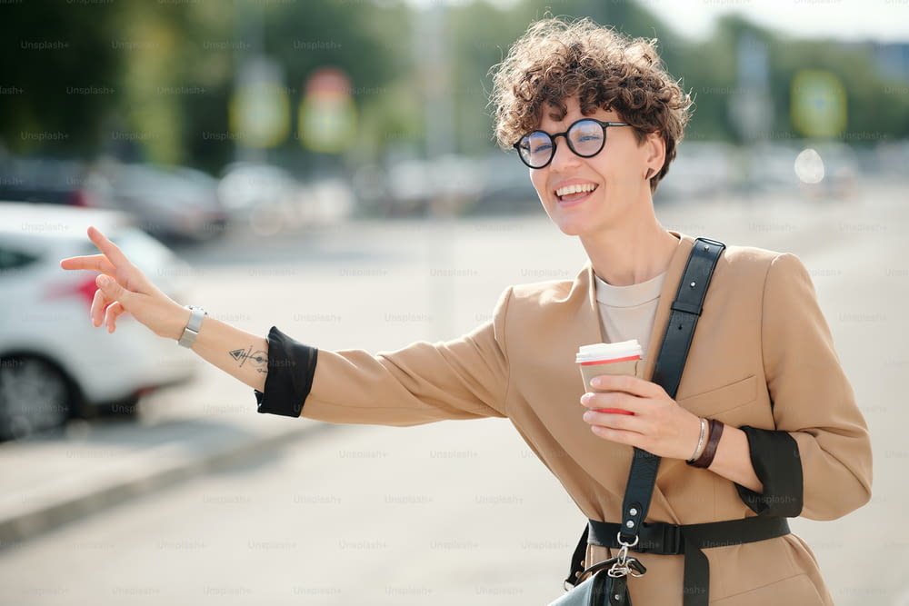 Young cheerful businesswoman with glass of coffee standing by road and catching taxi against modern building in urban environment