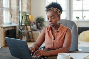 Seria giovane donna africana libera professionista in abbigliamento casual che lavora davanti al laptop mentre è seduta in poltrona vicino al tavolo nell'ambiente domestico