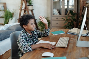オンラインレッスンの開始前に、コンピューターのモニターの前のテーブルに座って、先生に手を振る幸せな小さな男子生徒