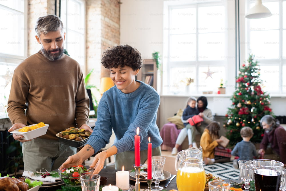 Feliz marido e mulher colocando salada caseira, batatas assadas, bebidas e outros alimentos na mesa festiva servida antes do jantar de Natal em família