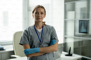 Portrait of smiling attractive nurse in blue latex gloves wearing badge on neck standing with crossed arms in office