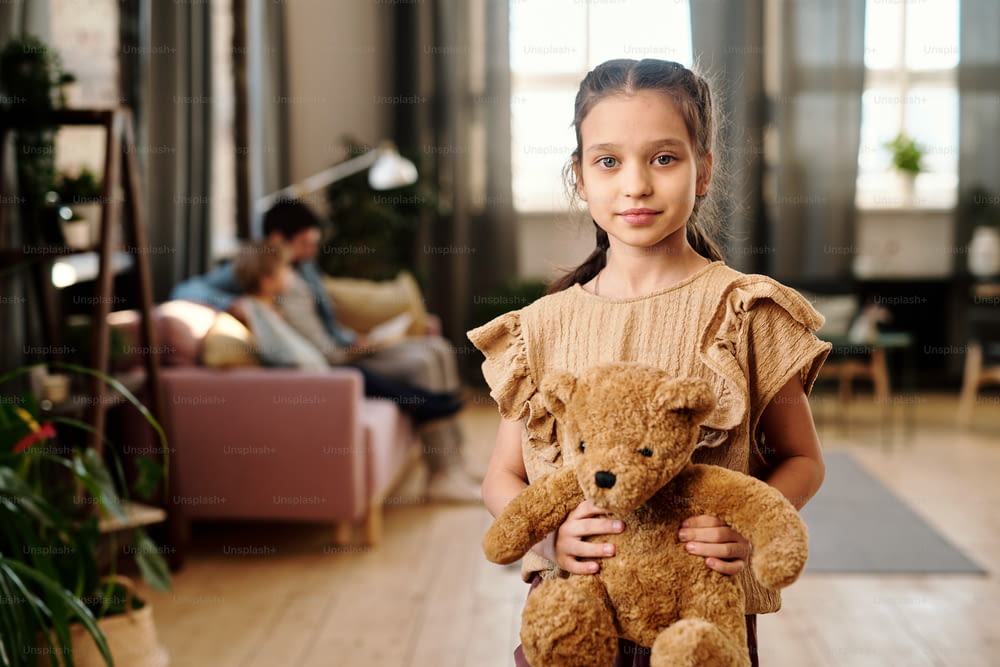 Cute girl with teddybear standing in living-room against her family