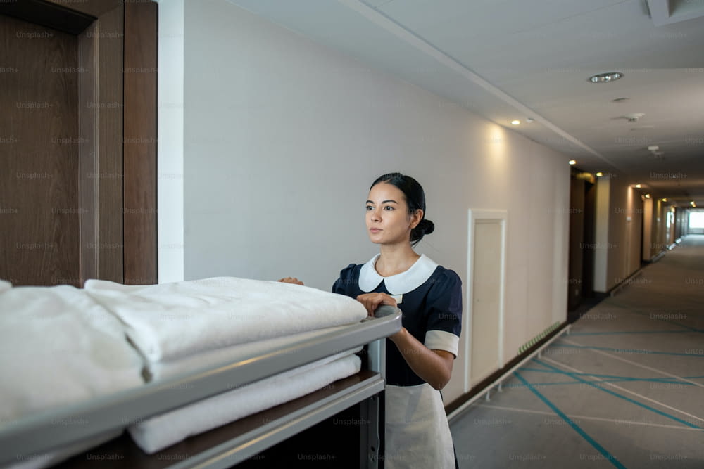 Jovem bela morena camareira ou trabalhador do hotel empurrando carrinho com toalhas limpas dobradas e outras coisas enquanto se move ao longo do corredor