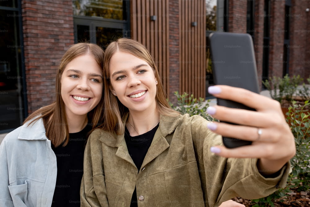 Duas meninas bonitas com cabelo loiro fazendo selfie contra o edifício moderno ao ar livre