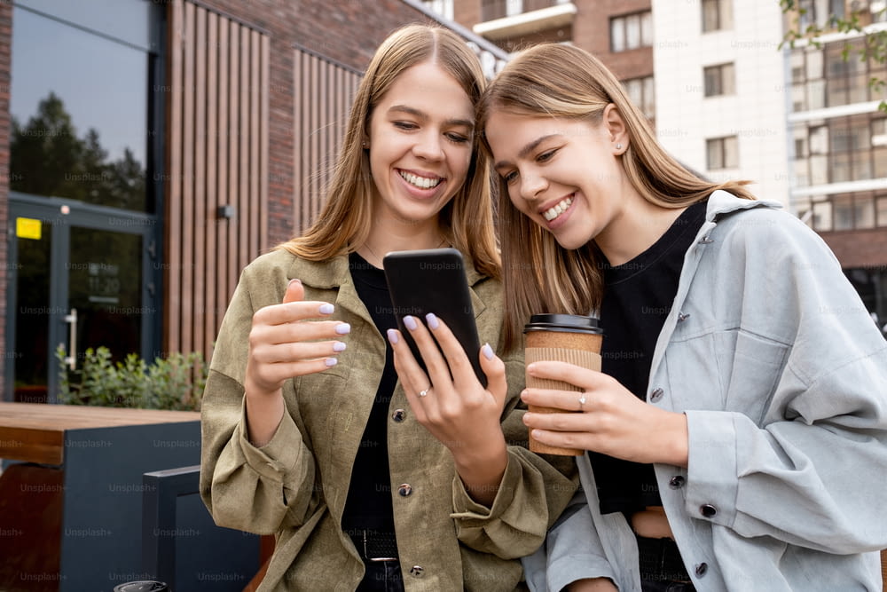 Young twin sisters in casualwear looking at smartphone screen while relaxing and having drinks in urban environment