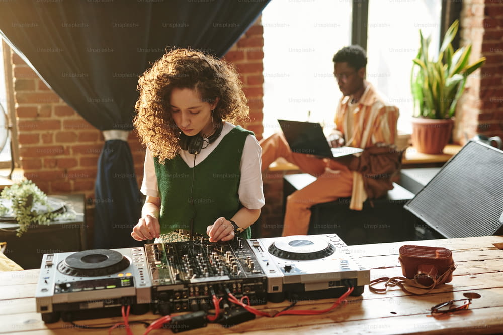 カジュアルウェアを着た若い女性が木のテーブルのそばに立ち、DJコントローラーを使いながら、黒人男性に対して余暇に新しい音楽を作る