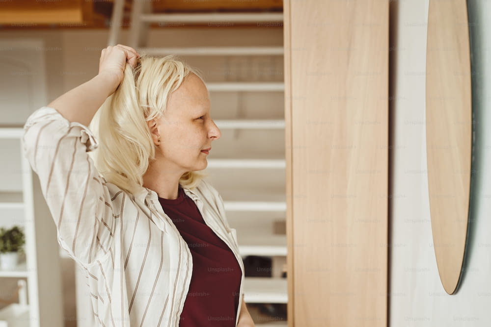 Warm-toned side view portrait of bald woman taking off wig while standing by mirror in home interior, alopecia and cancer awareness, copy space