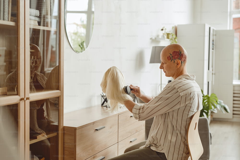 Side view portrait of modern bald woman brushing wig while sitting by mirror in home interior, alopecia and cancer awareness, copy space