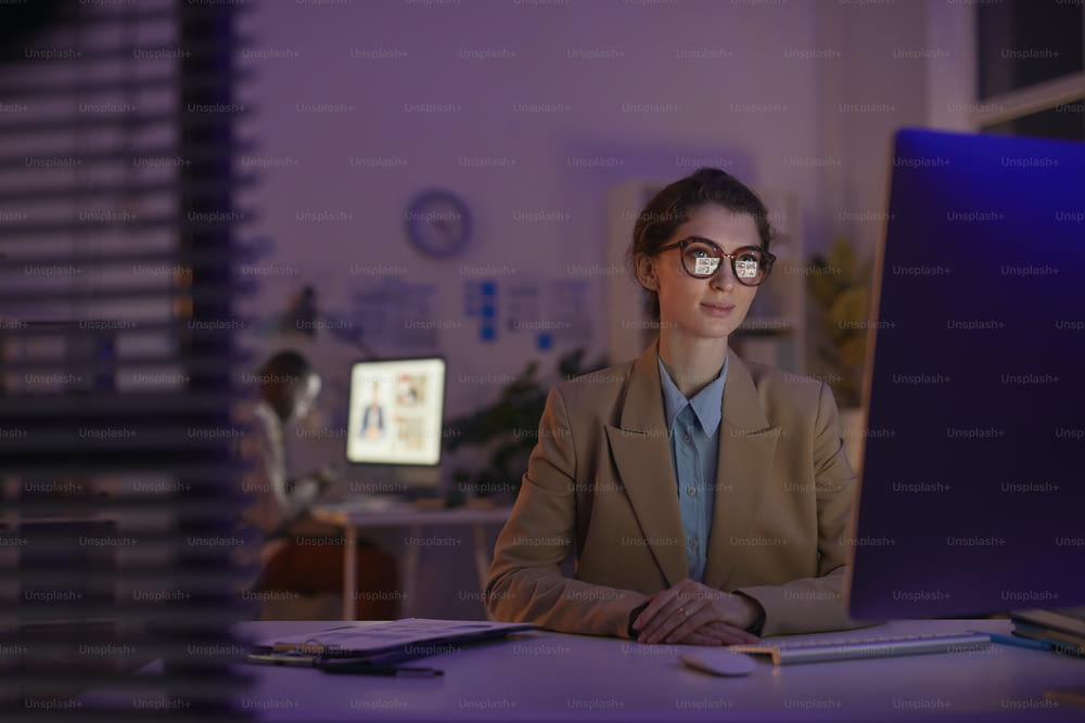 Beautiful young woman wearing eyeglasses sitting at office desk late in evening watching something on computer monitor