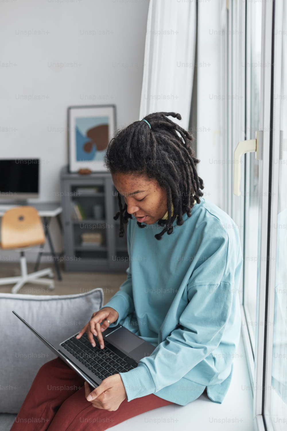 Vertical portrait of real African American woman using laptop while sitting on by window at home with focus on skin imperfections