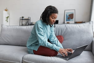 Retrato de cuerpo entero de una mujer afroamericana real usando una computadora portátil mientras está sentada en el sofá de su casa con enfoque en las imperfecciones de la piel, espacio de copia