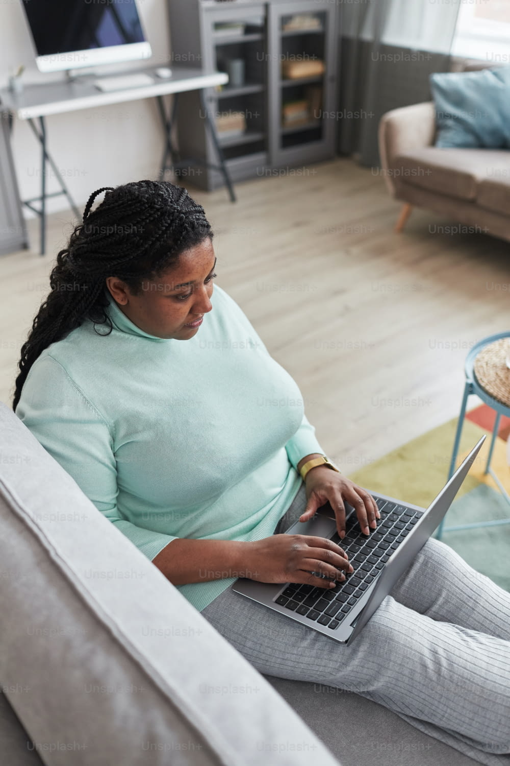 Vertical portrait of curvy African American woman using laptop while enjoying work from home relaxing on couch in minimal grey interior