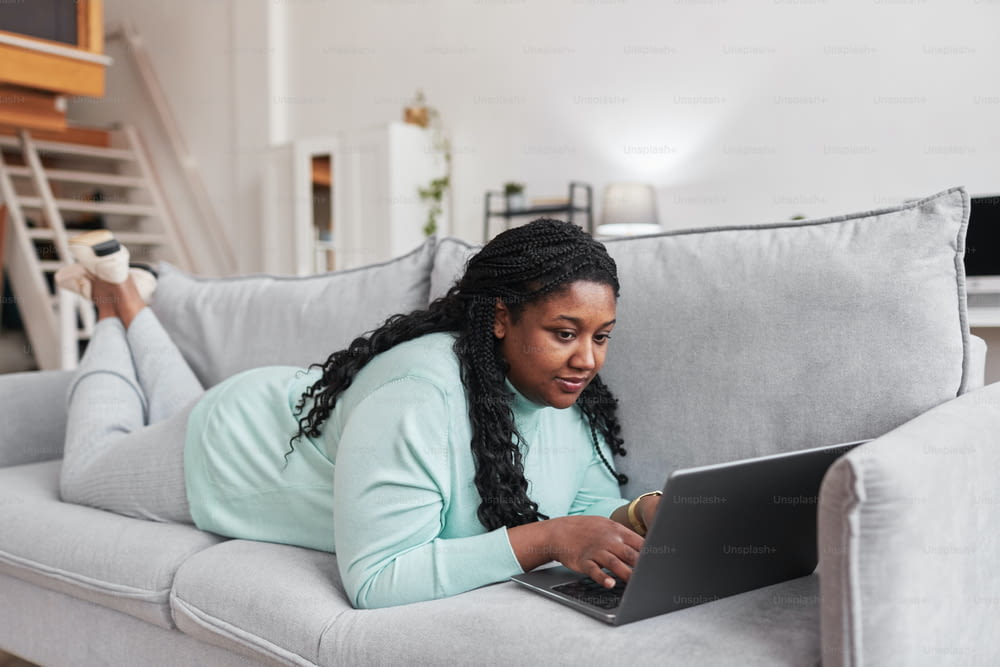 Full length portrait of curvy African American woman using laptop while lying on couch and relaxing at home in minimal interior