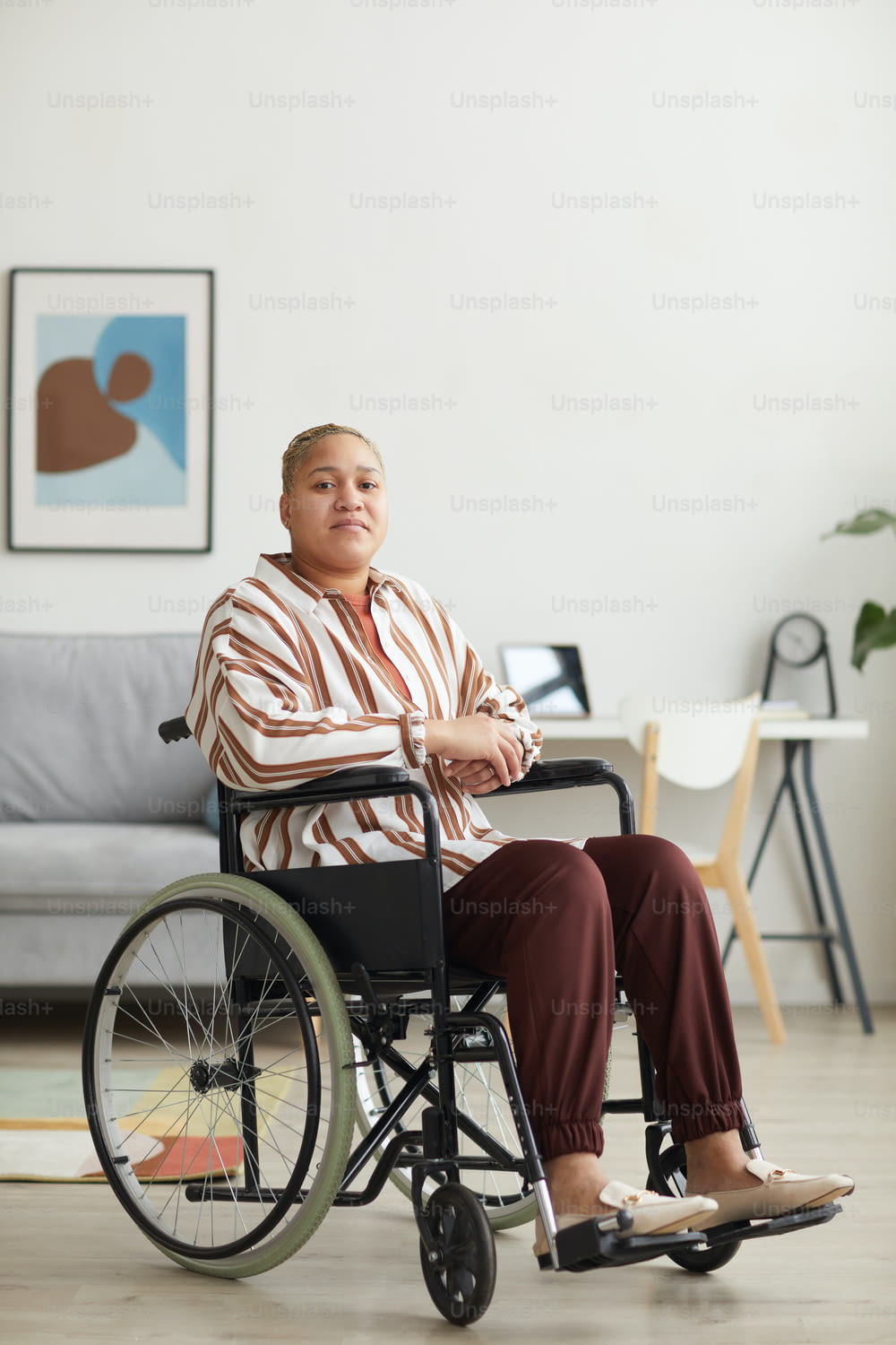 Retrato vertical de corpo inteiro da mulher moderna mestiça sentada em cadeira de rodas e olhando para a câmera no interior mínimo da casa, espaço de cópia