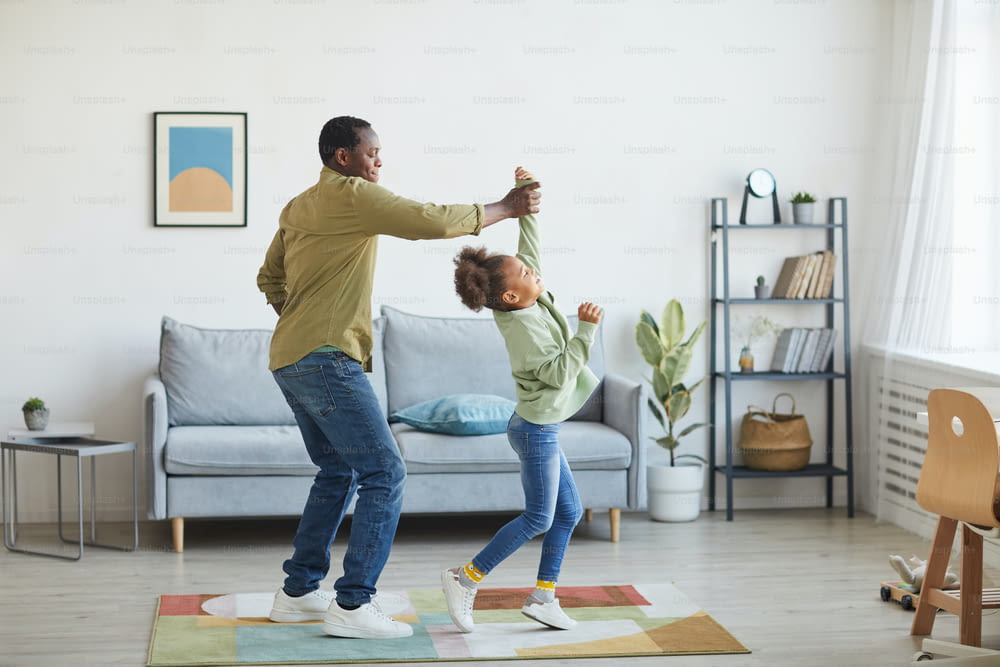Full length portrait of happy African-American father dancing with daughter while having fun in minimal home interior, copy space