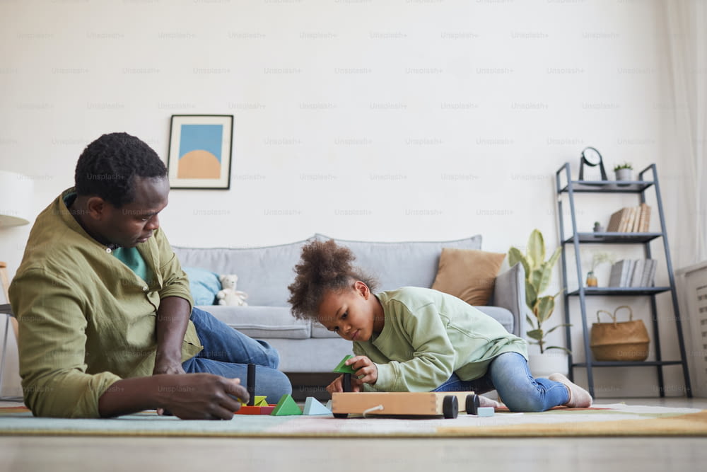 Side view portrait of African-American man playing with daughter while sitting on floor in cozy home interior, copy space
