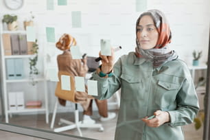 Waist up portrait of modern Middle-Eastern woman working in office and placing sticker notes on glass wall, copy space