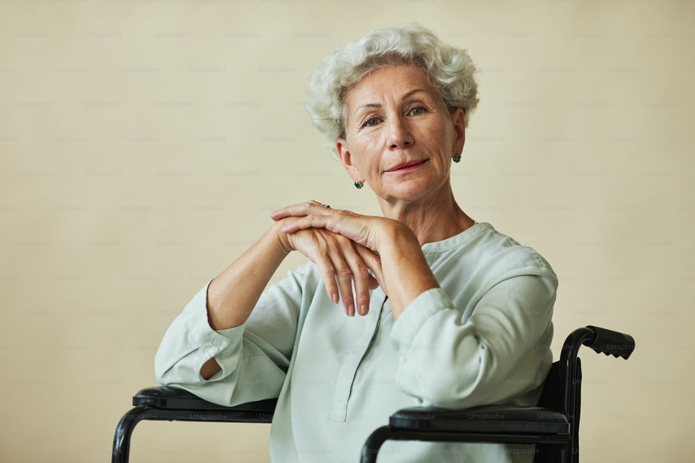 Porträt einer eleganten Seniorin, die im Rollstuhl vor neutralem Hintergrund in die Kamera schaut, Kopierraum