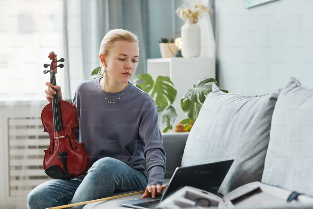 Retrato de una joven rubia tocando el violín en casa y viendo una lección en línea, espacio de copia